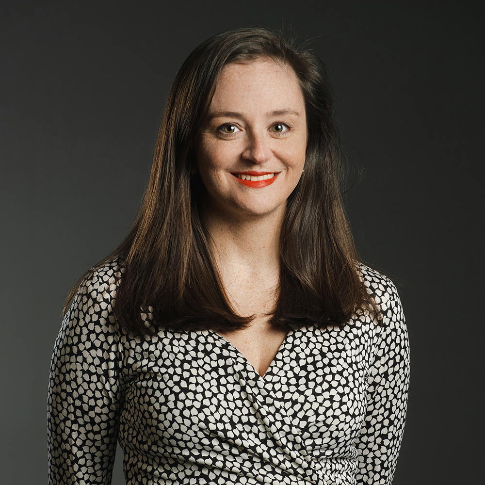 Anne L. Williams Color photograph of a light skinned woman with long brown hair wearing a black and white patterned dress and smiling at the camera