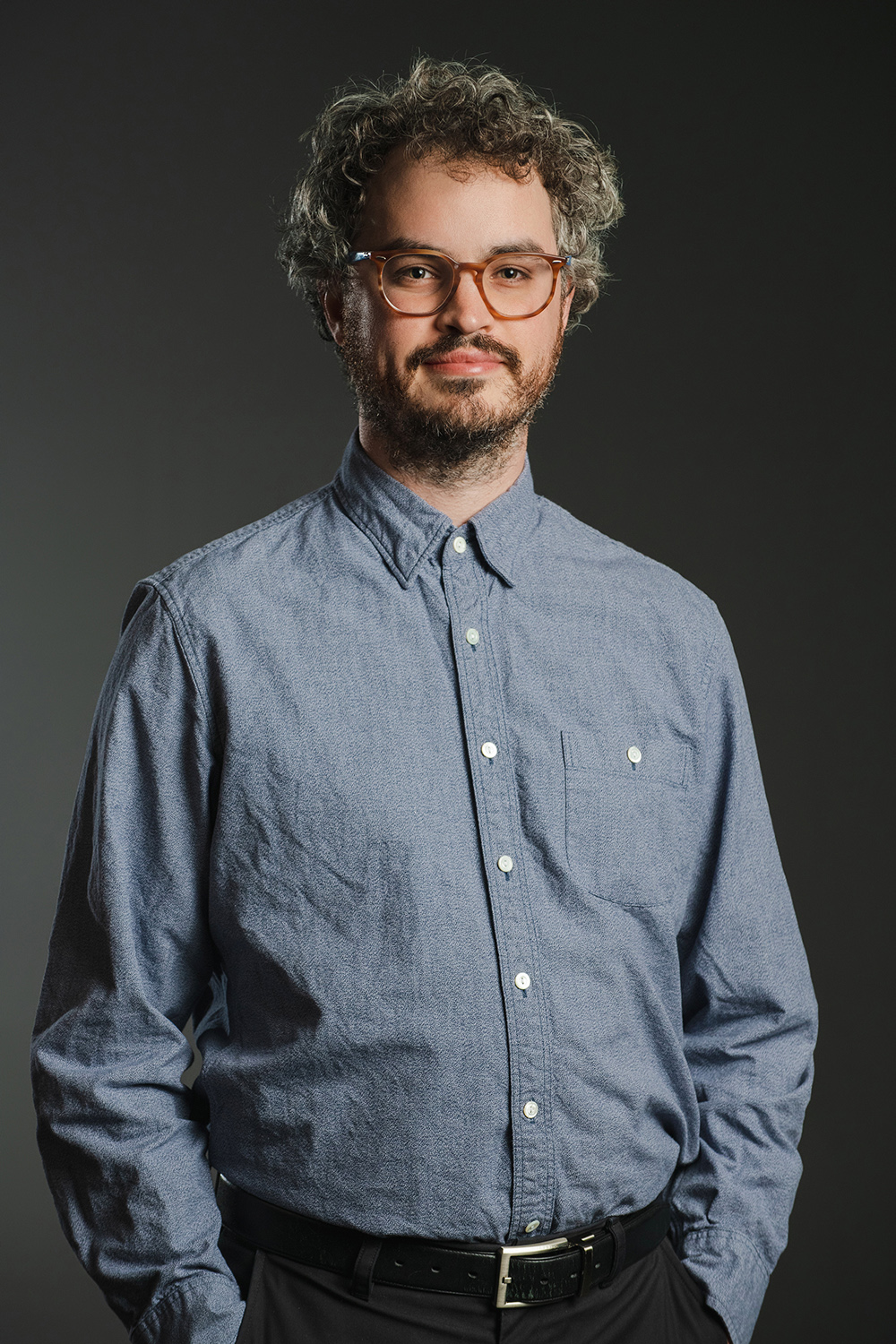 Anthony Vine Color photograph of a light skinned man wearing glasses a blue button-up shirt; he looks at the camera with his hands in his pockets