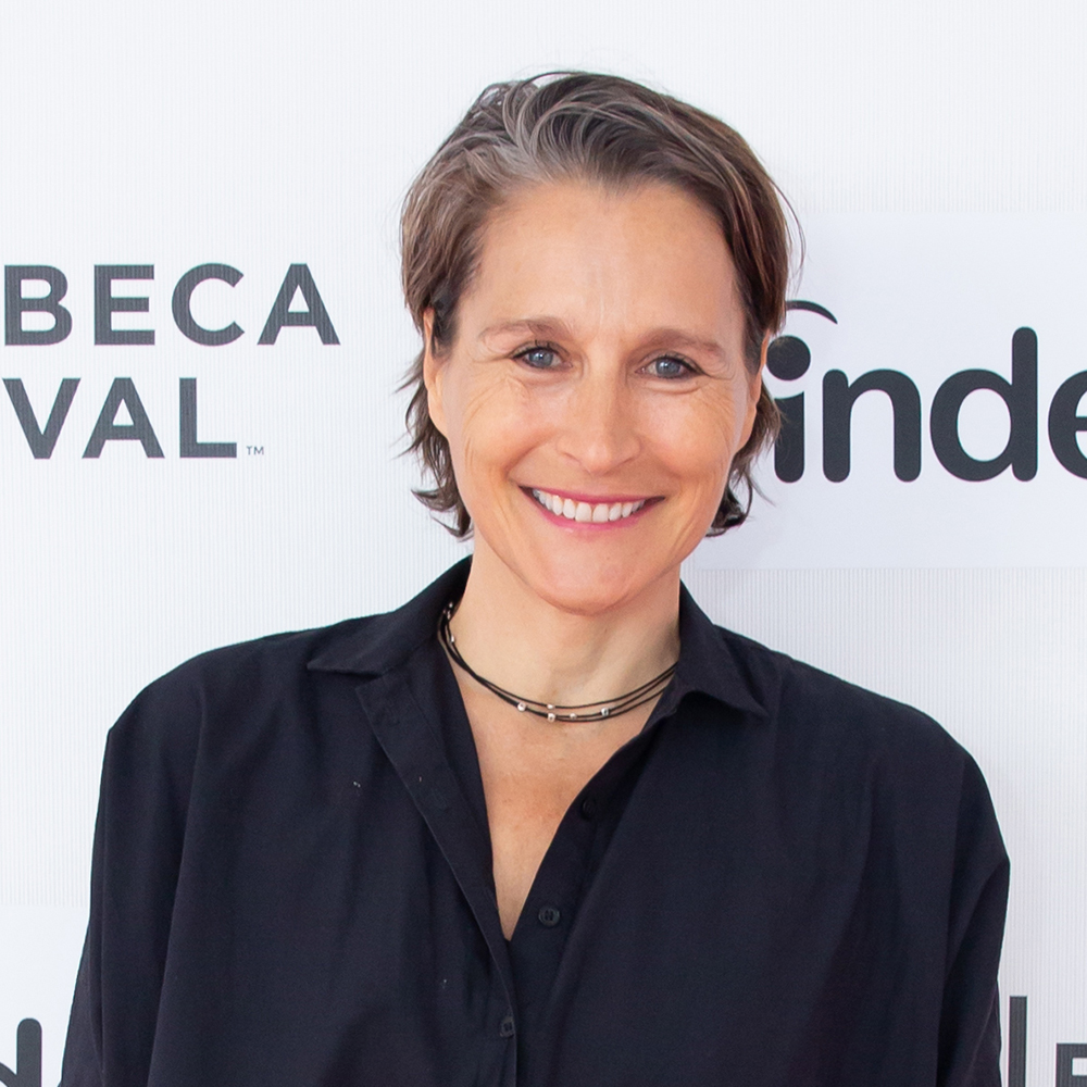Catherine Gund Color photo of the head and shoulders of a light skinned woman wearing a dark blue top; she stands in front of a step and repeat and smiles at the camera
