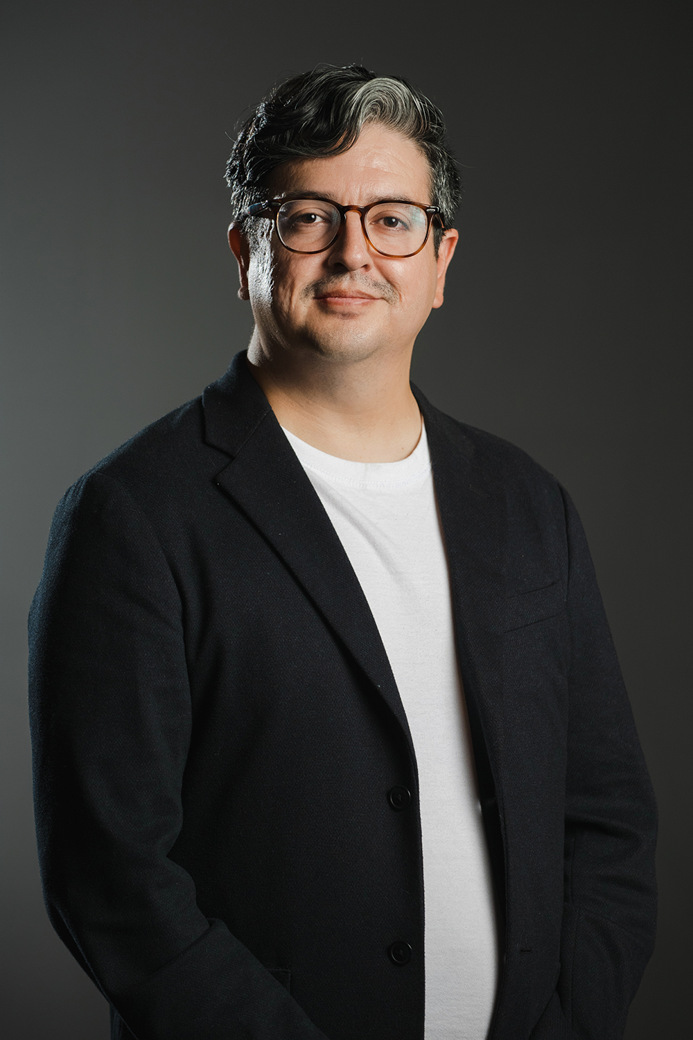 César A. Lopez Color photograph of a man wearing glasses and a dark blazer smiling slightly at the camera