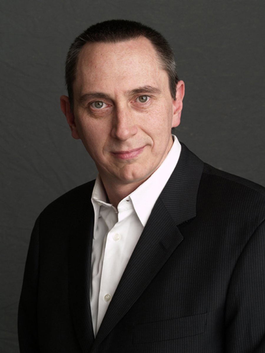 Color photograph of the head and torso of a light skinned man wearing a dark suit over a white shirt