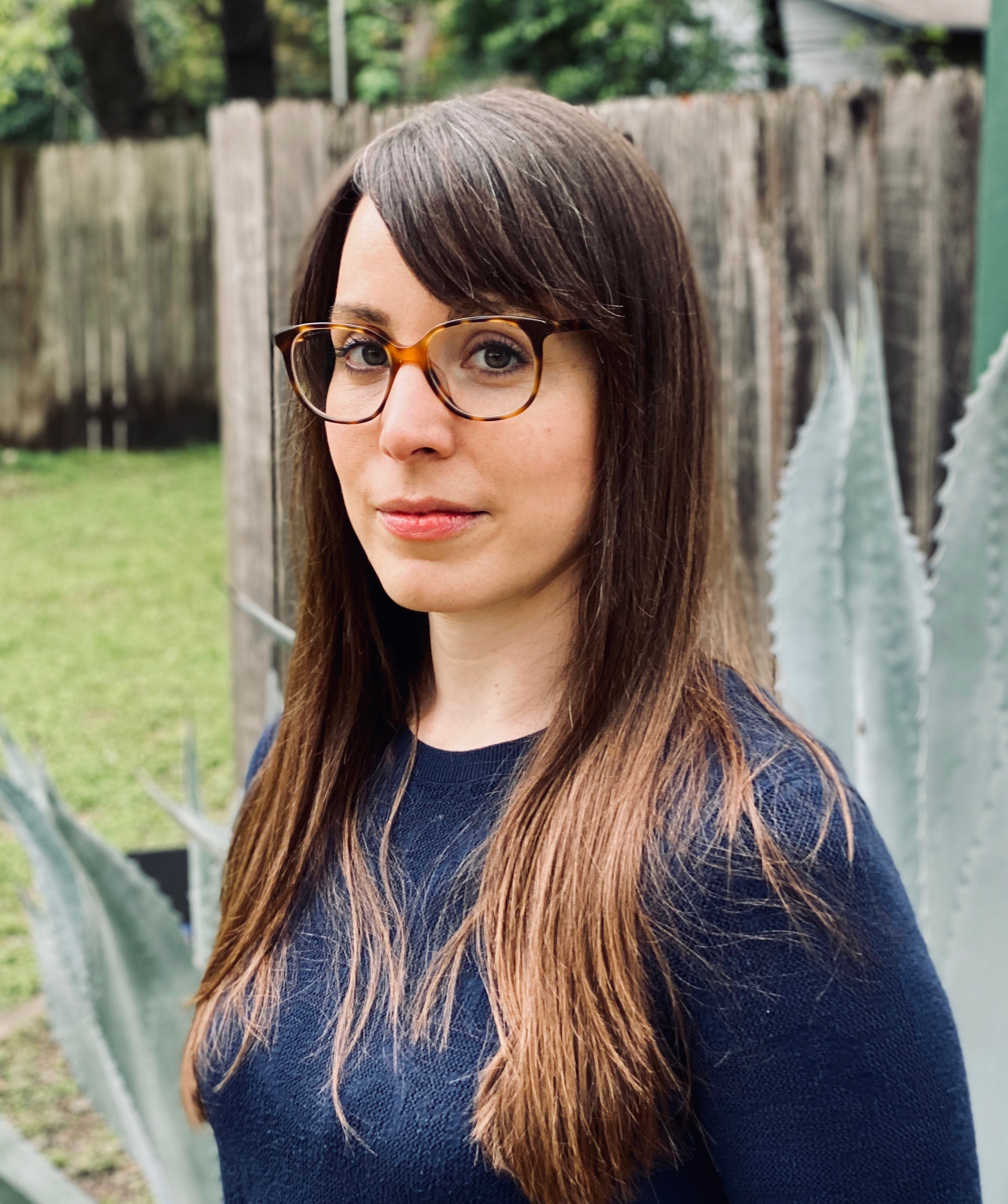 Christy Q. Schirmer Color photographic portrait of Christy Q. Schirmer with glasses and a blue top, with a large aloe plant and a wooden fence behind her