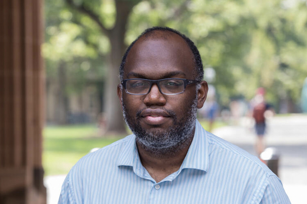 Dan-el Padilla Peralta Color photograph of the head and shoulders of a dark skinned mean with a salt and pepper beard wearing glasses and a light blue open collar shirt; he looks at the camera in a serious way