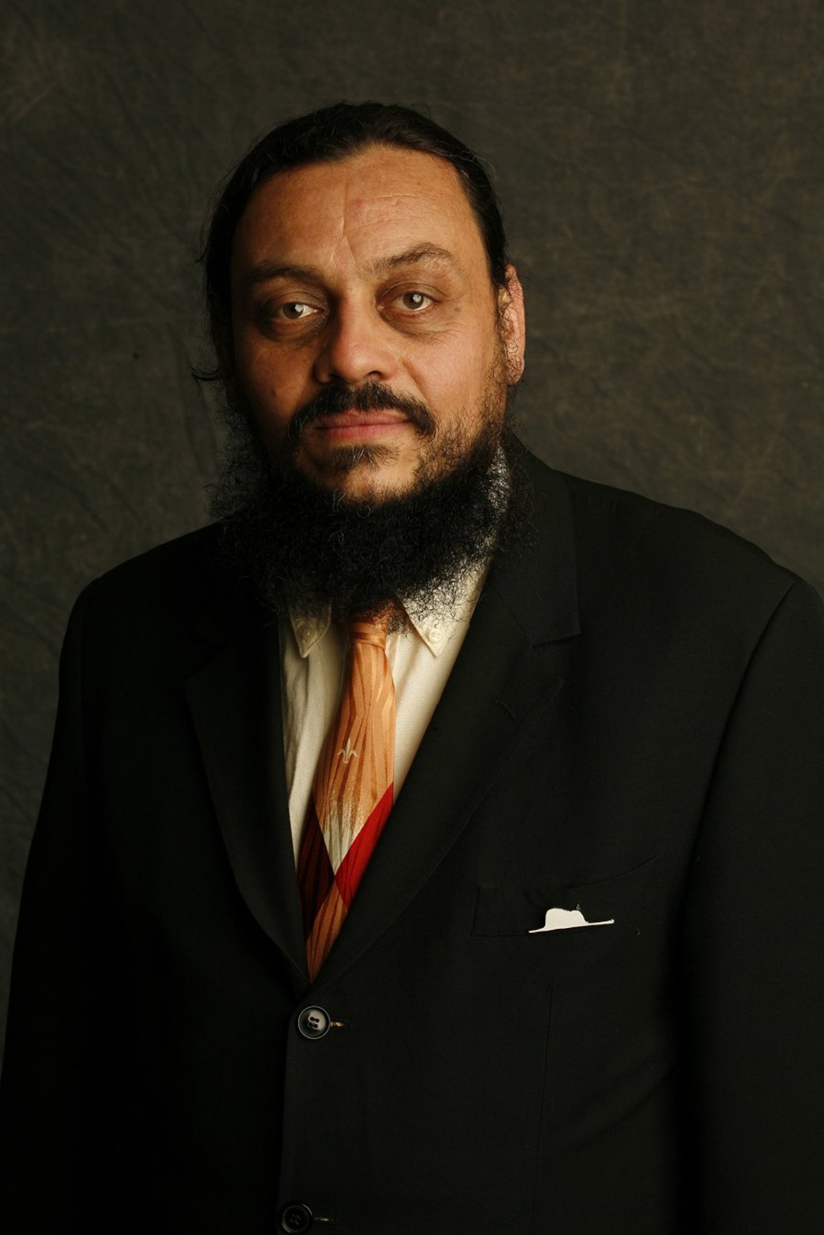 Color photograph of the head and torso of a olive skinned with a beard wearing a suit and tie and looking at the camera