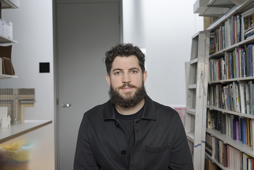 David Costanza Color photo of a man looking at the camera; he is in a room with bookshelves, ladder, door, and desk ledge