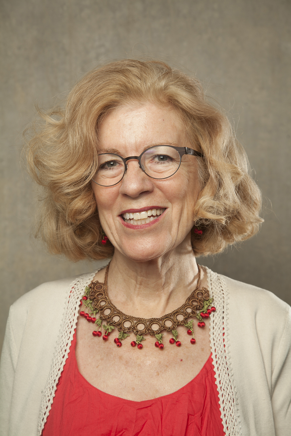 Color photograph of Diane Favro, a woman wearing glasses, a red top, and tan sweater, looking straight at the camera and smiling