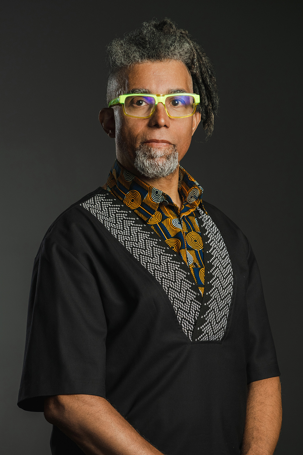 Dread Scott Color photograph of a dark skinned man with gray dreadlocks and goatee wearing glasses and an African inspired shirt, looking directly at the camera