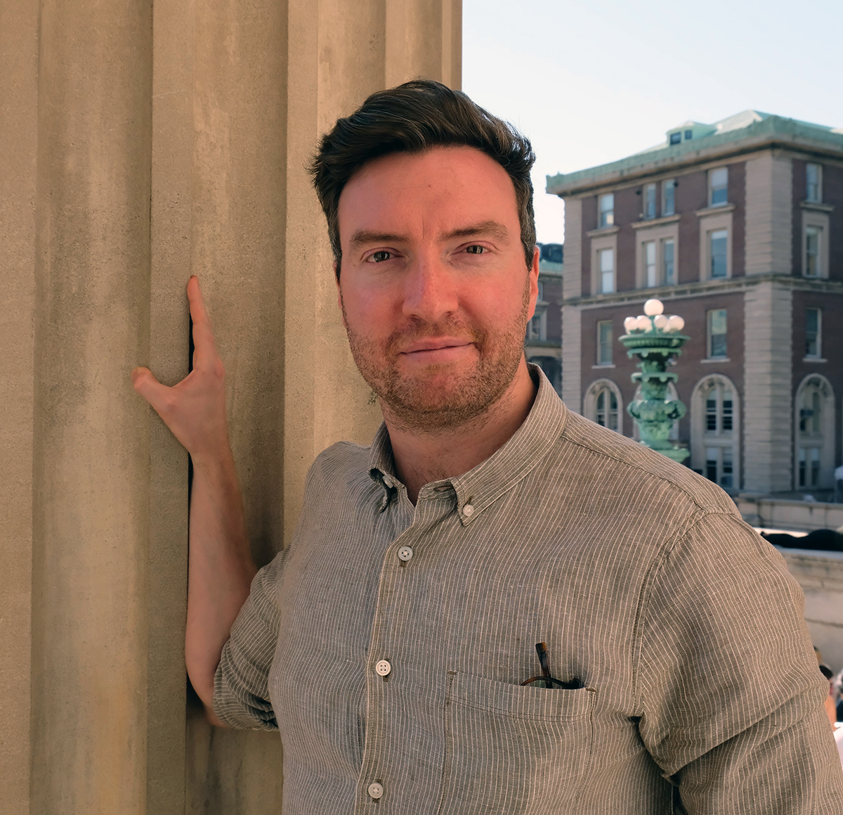 Evan Jewell Color photograph of a light skinned man with brown hair and a gray button-up shirt standing next to a fluted stone column, on which his hand rests; he looks directly at the camera