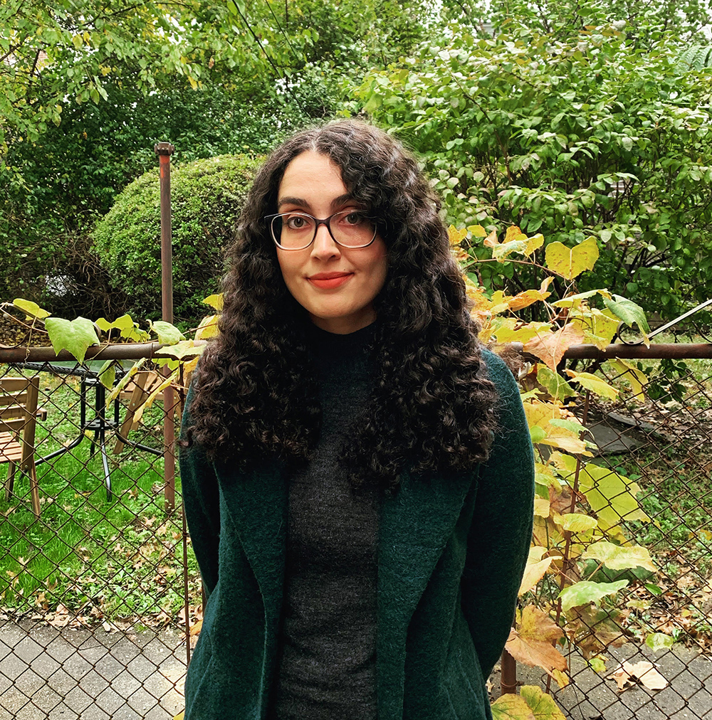 Gabriella L. Johnson Color photograph of a light skinned woman with dark curly hair wearing glasses and dark clothes; behind her is a chain linked fence, patio furniture, and green shrubs and trees