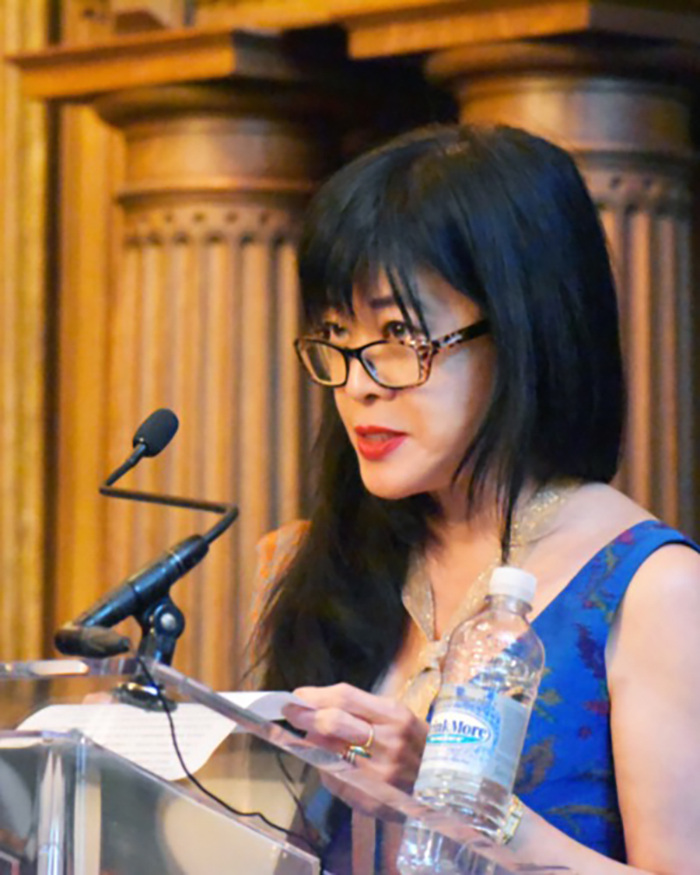 Geraldine Heng Color photo of an Asian woman wearing a blue dress and glasses and standing at a podium with a microphone