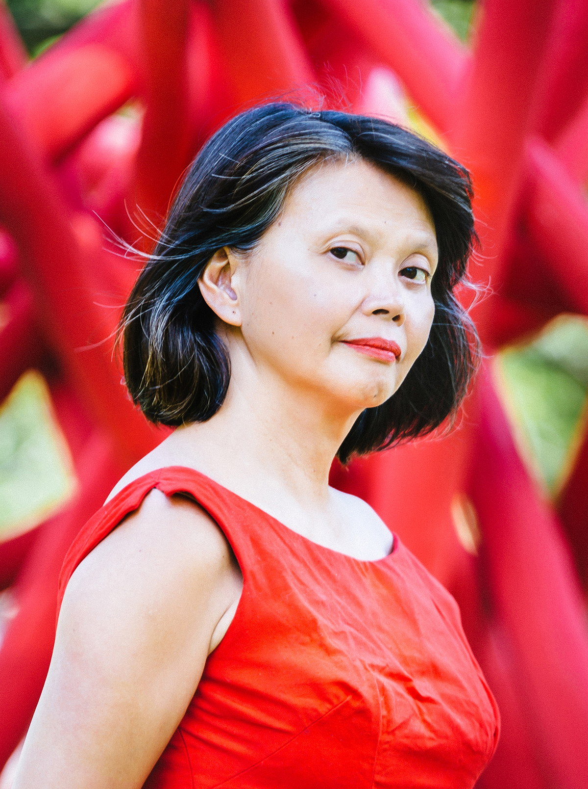 Gina Apostol Color photograph of the head and torso of a light skinned Filipino woman with dark hair, wearing a wrinkled red dress and looking at the camera; she stands in front of what looks like a very large but blurred foliage