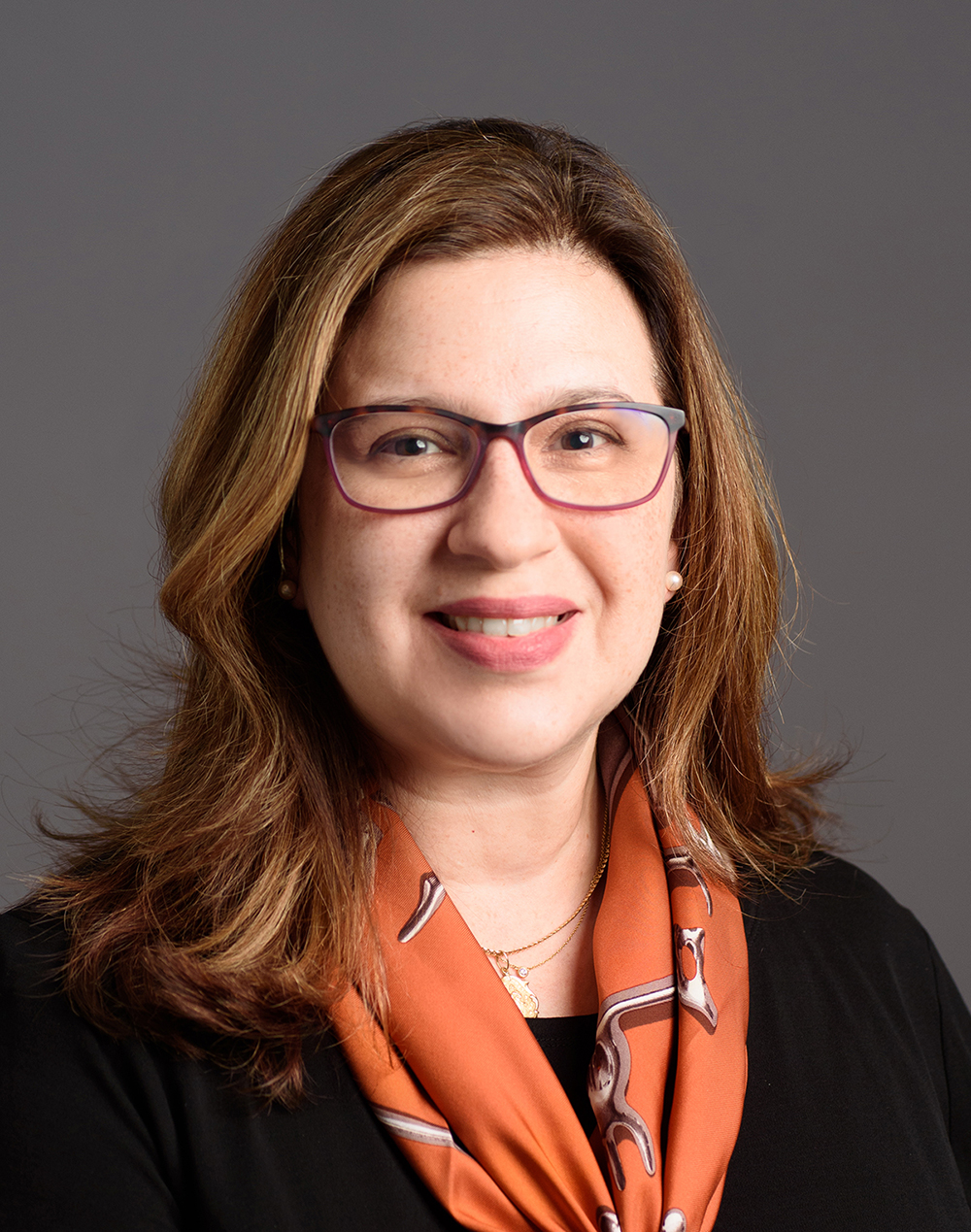 Hérica Valladares Color photograph of the head and shoulders of a light skinned woman with brown hair smiling at the camera; she wears glasses and a patterned orange-clay scarf