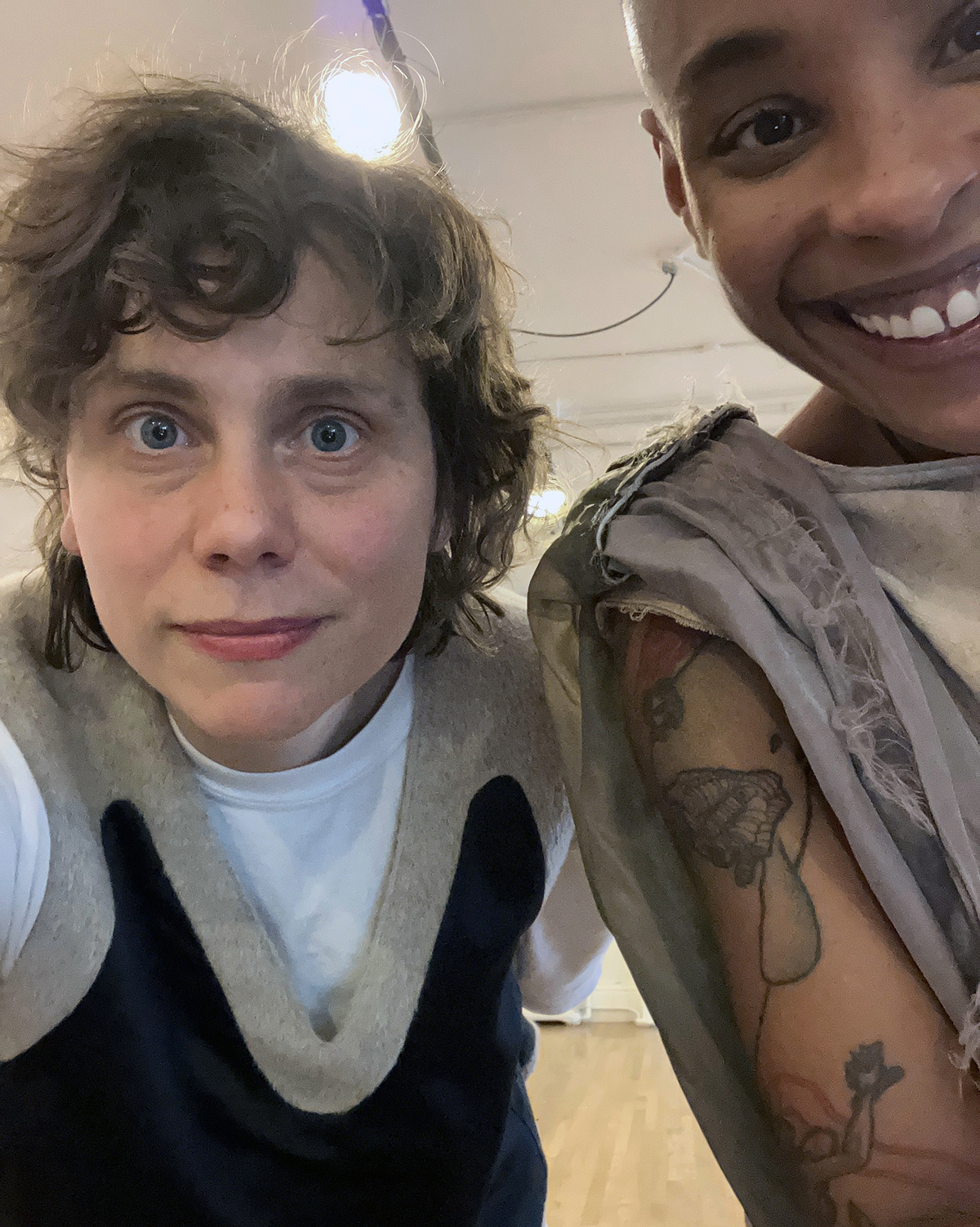 Jasmine Hearn and Athena Kokoronis Color photograph of two women filling the frame: on the left is a light skinned woman staring at the camera; on the right is a dark skinned woman smiling at the camera while wearing a sleeveless top, showing her arm tattoes