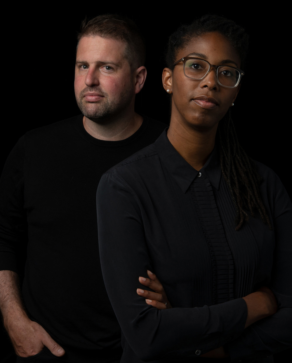 Jennifer Newsom and Tom Carruthers Color photograph of the head and torsos of two people: on the left is a light skinned man with his head turned slightly but still looking at the camera with one hand in his pocket; on the right is a dark skinned woman wearing glasses and looking at the camera with her arms folded across her chest; both are wearing black clothes and stand against a black background