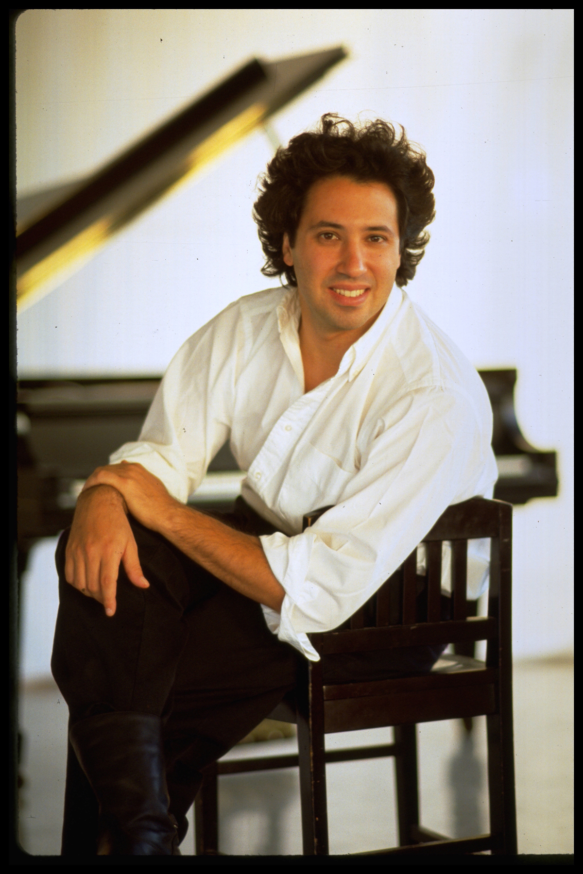 John Davis Color photograph of a light skinned man with brown hair wearing a white button-up shirt and dark trousers and smiling at the camera; he sits on a chair and behind him a piano with an open lid can be seen