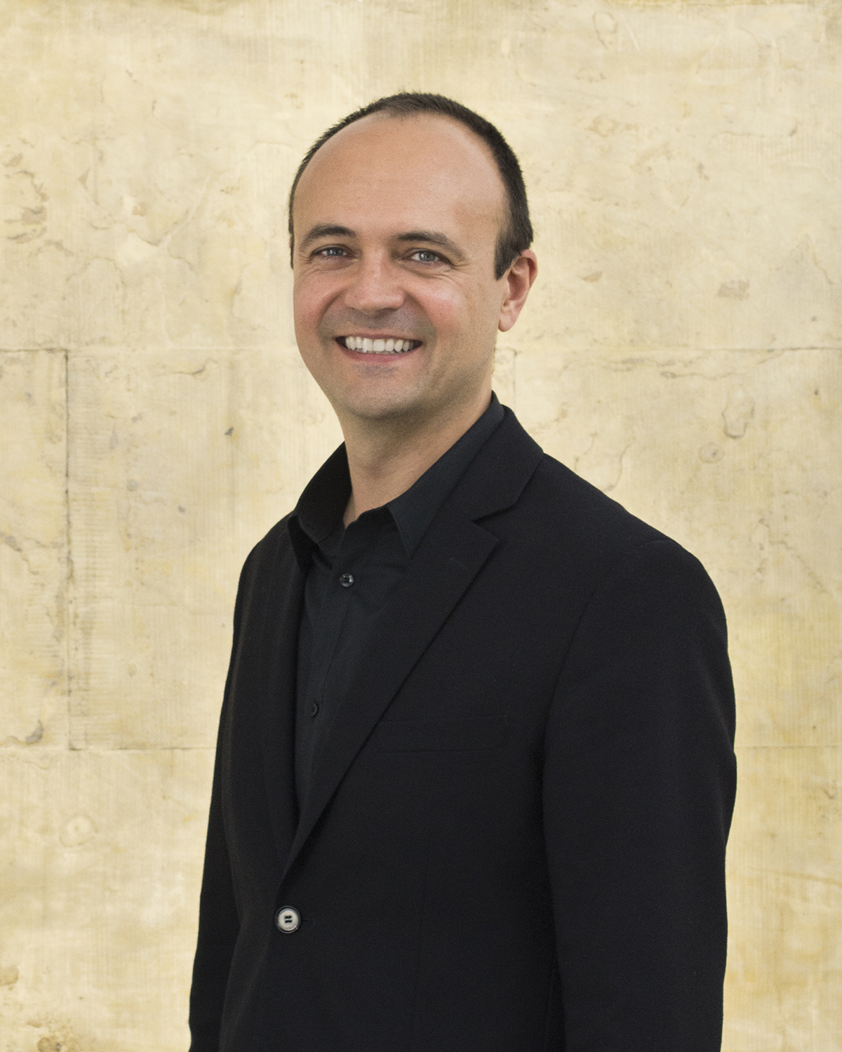 Jorge Otero-Pailos Color photograph of the Jorge Otero-Pailos from the waist up; he is light skinned, has closely cropped fair, wears a black shirt and blazer, and smiles directly at the camera