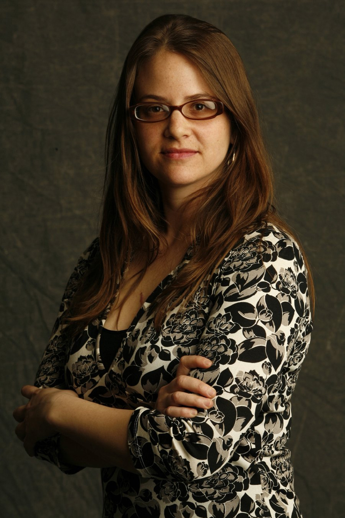 Kate Gilmore Color photograph of the head and torso of a light skinned woman wearing a black and white patterned top and glasses; she crosses her arms as she looks at the camera