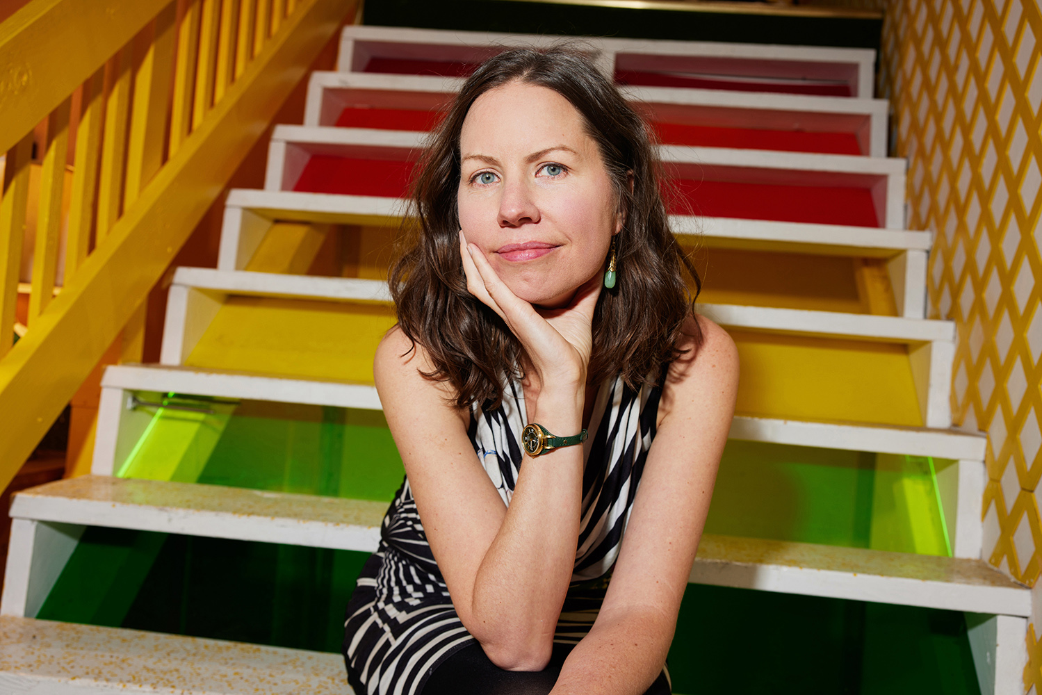 Kate Soper Color photograph of a light skinned woman sitting on a multicolored staircase; she looks at the camera as she rests her head on her hand
