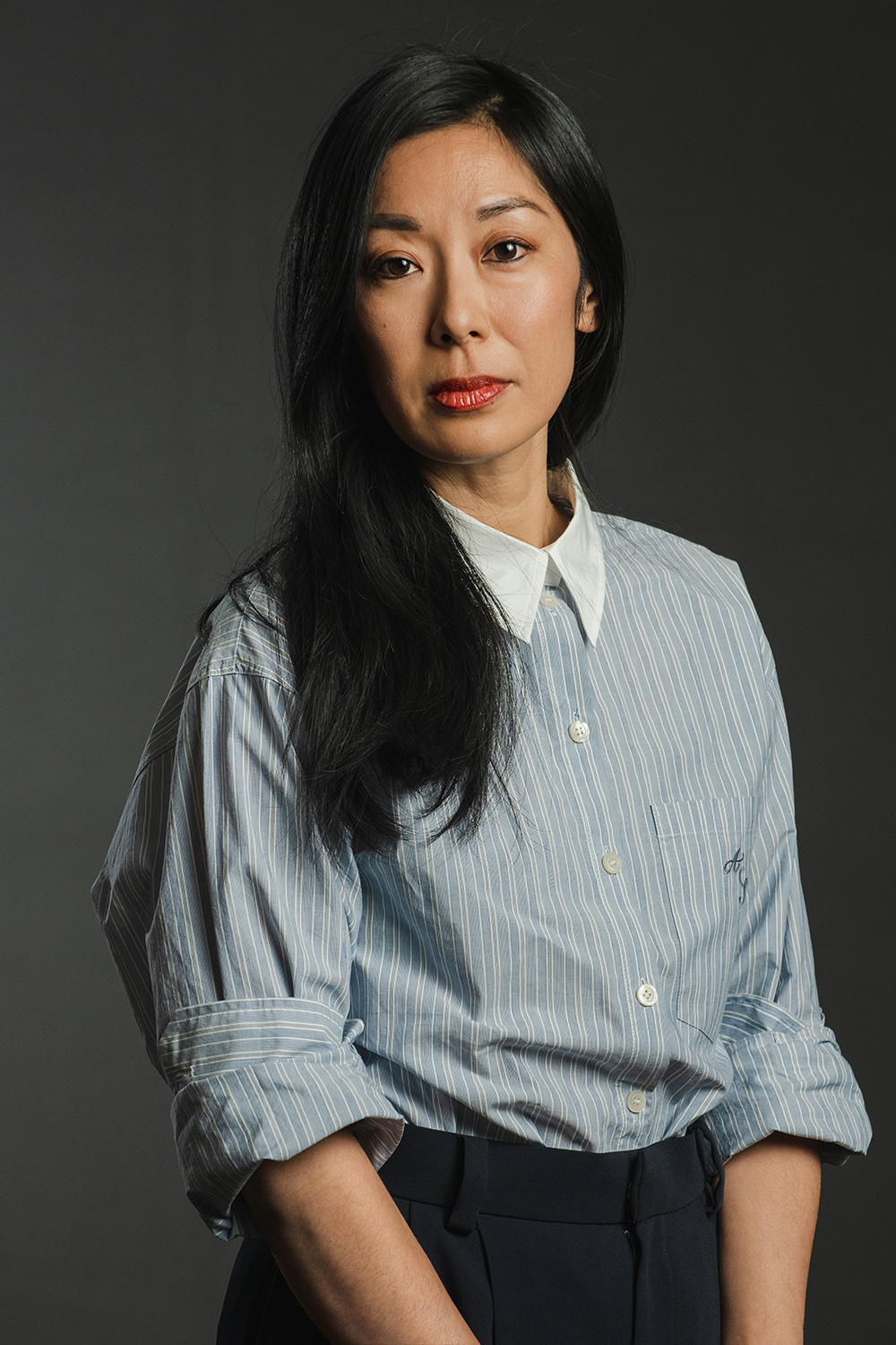 Katie Kitamura Color photograph of an Asian woman with long dark hair and a blue striped button up top with sleeves rolled, looking directly at the camera with a serious expression