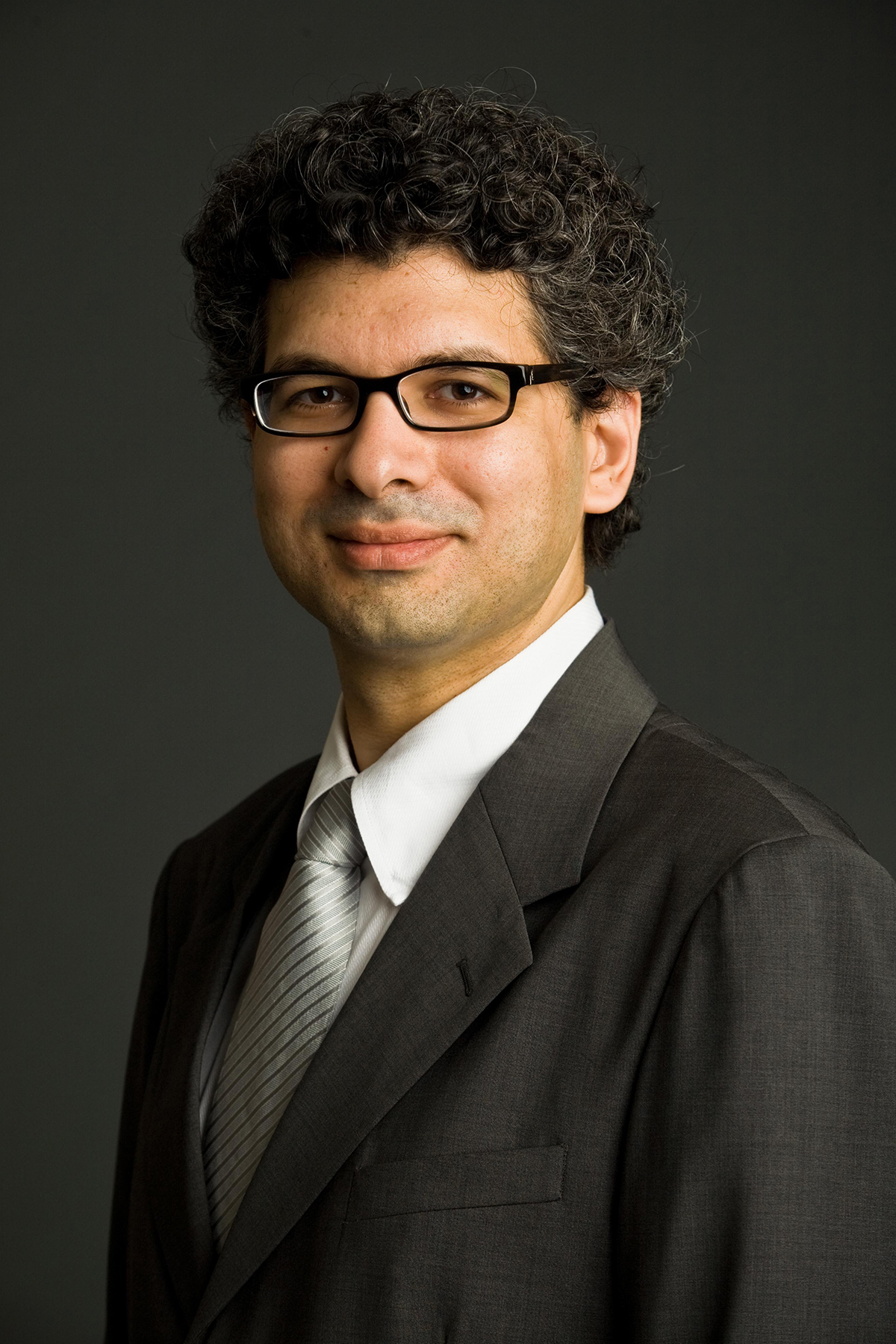 Keeril Makan Color photograph of a light skinned man wearing glasses, a suit, and a tie in a photographer's studio, smiling at the camera