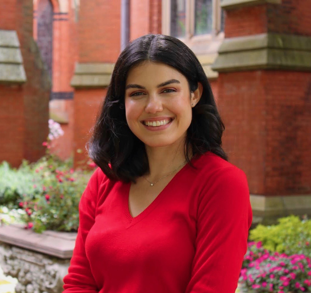 Mary C. Danisi Color photograph of a medium skinned woman with dark hair wearing a bright red sweater; she is outdoors in front of a brick building, smiling at the camera