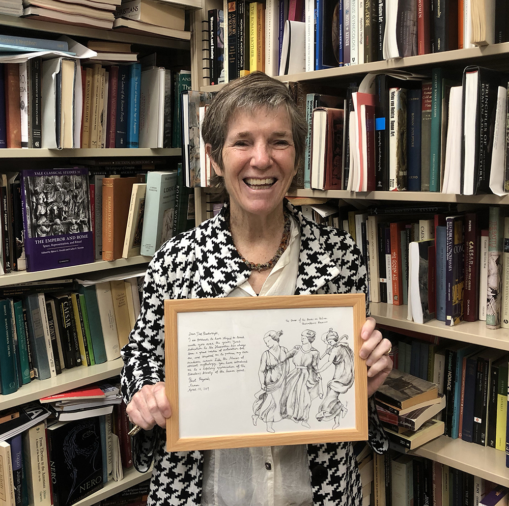Color photograph of Mary Tolly Boatwright standing in front of book-filled shelves and holding a framed handwritten letter and drawing