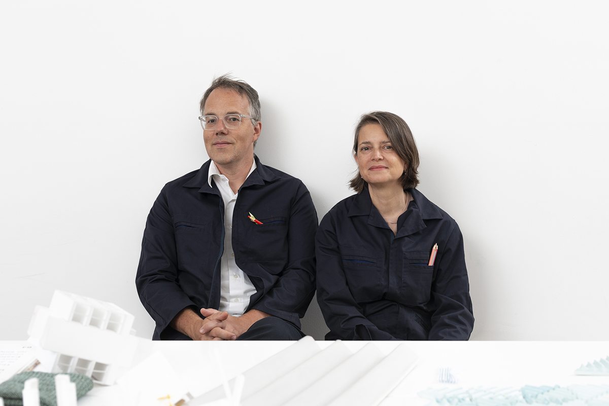 Michael Meredith and Hilary Sample Color photograph of two light skinned people (a man and a woman) smiling slightly at the camera as they sit behind a white table and in front of a white wall wearing matching navy jumpsuits with colored pencils in their shirt pockets (his jumpsuit is open at the chest, revealing a white button-up shirt underneath)