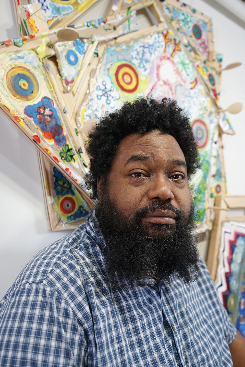 Mike Cloud Color photograph of a dark skinned man with a long dark bear and wearing a checkered blue and white shirt; he sits underneath a colorful abstract artwork and looks directly at the camera