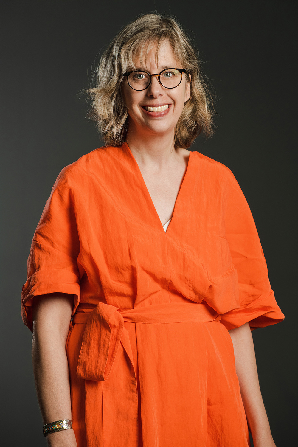Miranda E. Mote Color photograph of a light skinned woman with wavy blond hair wearing glasses and an orange jumpsuit