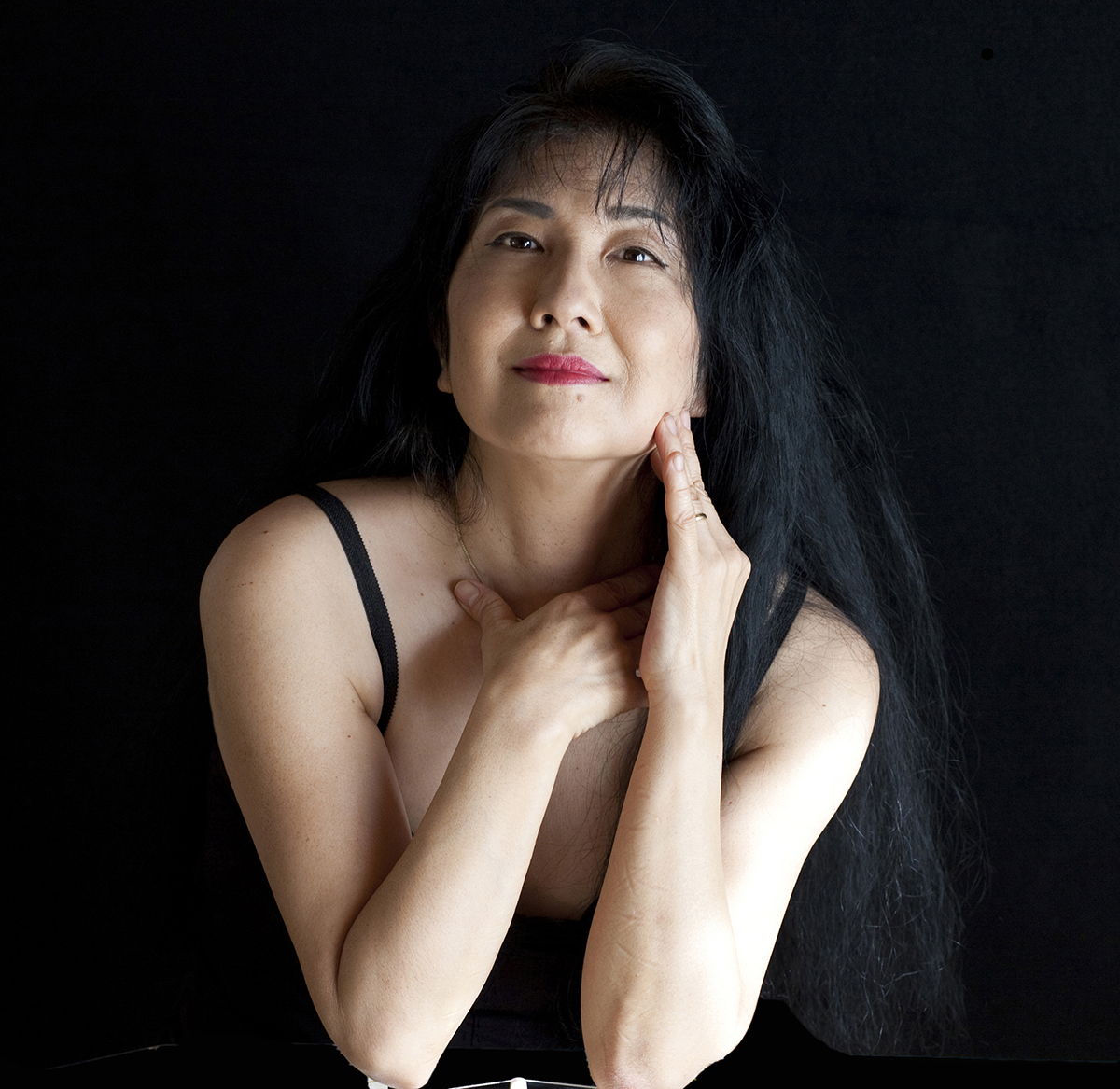 Miya Masaoka Color photograph of an Asian woman with dark hair wearing a black thin strapped dress and smiling at the camera; her left hand touches her cheek, and her right hand touches her throat