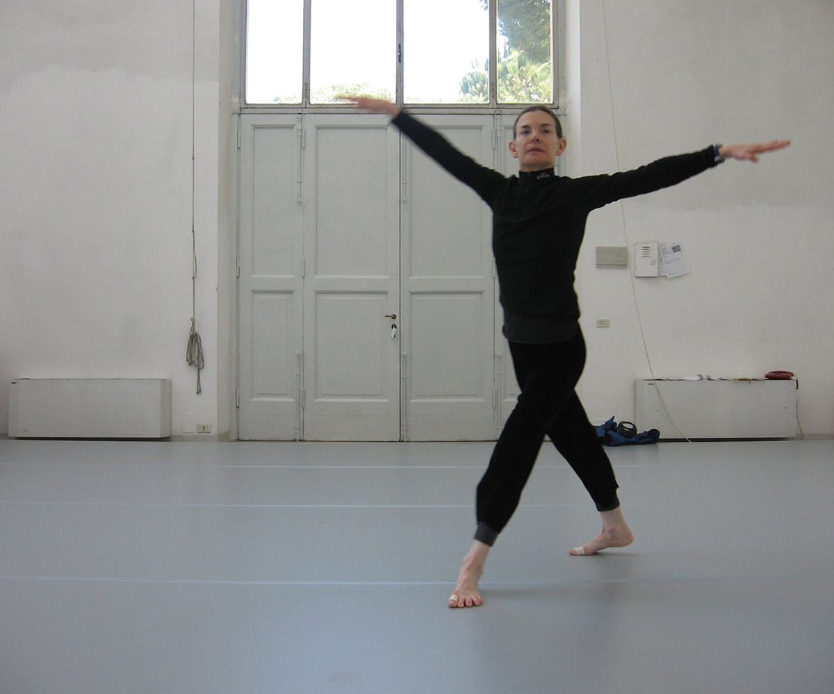 Molissa Fenley Color photograph of a light skinned woman in a dark bodysuit dancing in a white-walled studio with her arms and legs outstretched