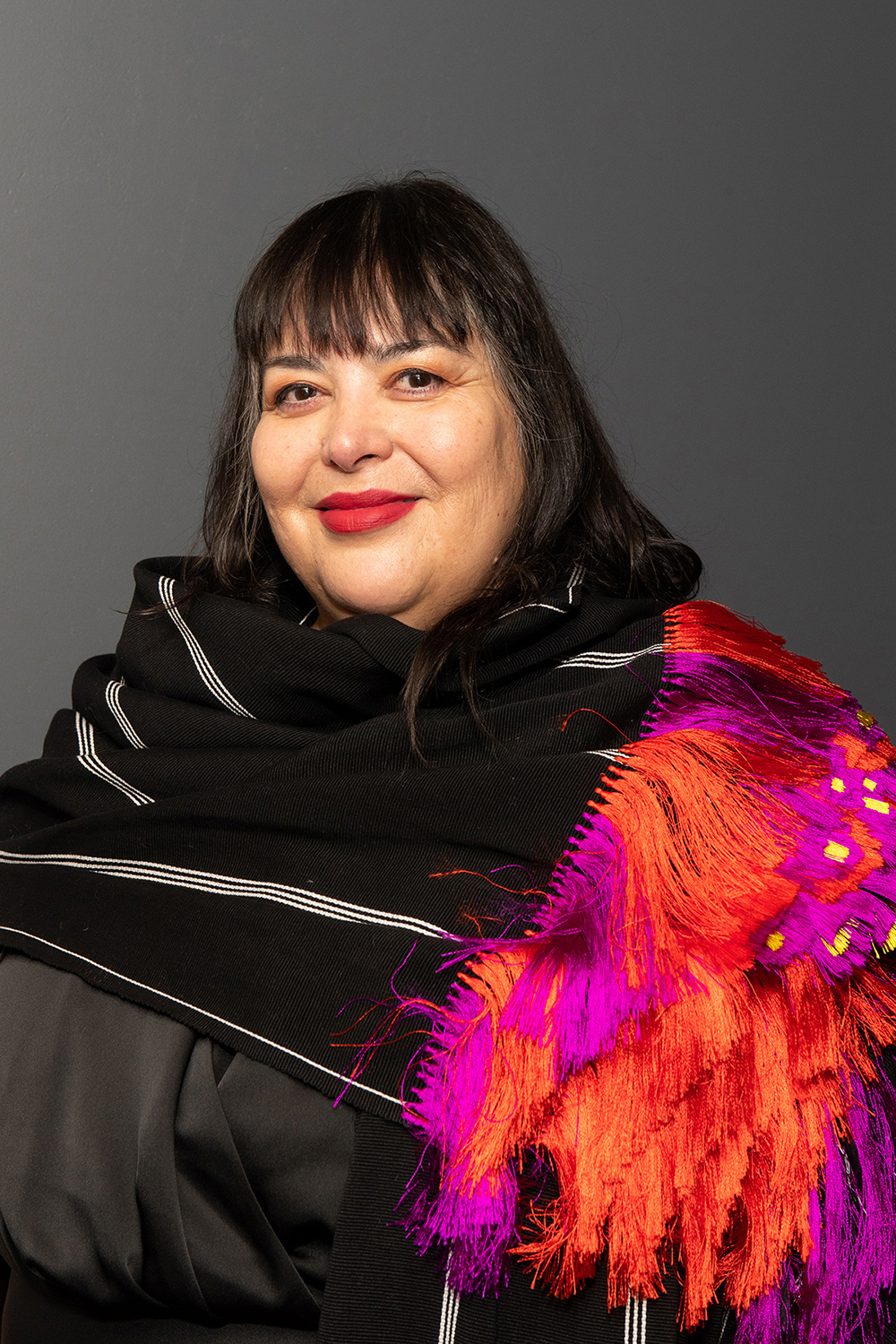 Nao Bustamante Color photograph of a light skinned woman with dark hair and bright red lipstick smiling at the camera; she wears a complex, multilayered garment in black, fucsia, and orange