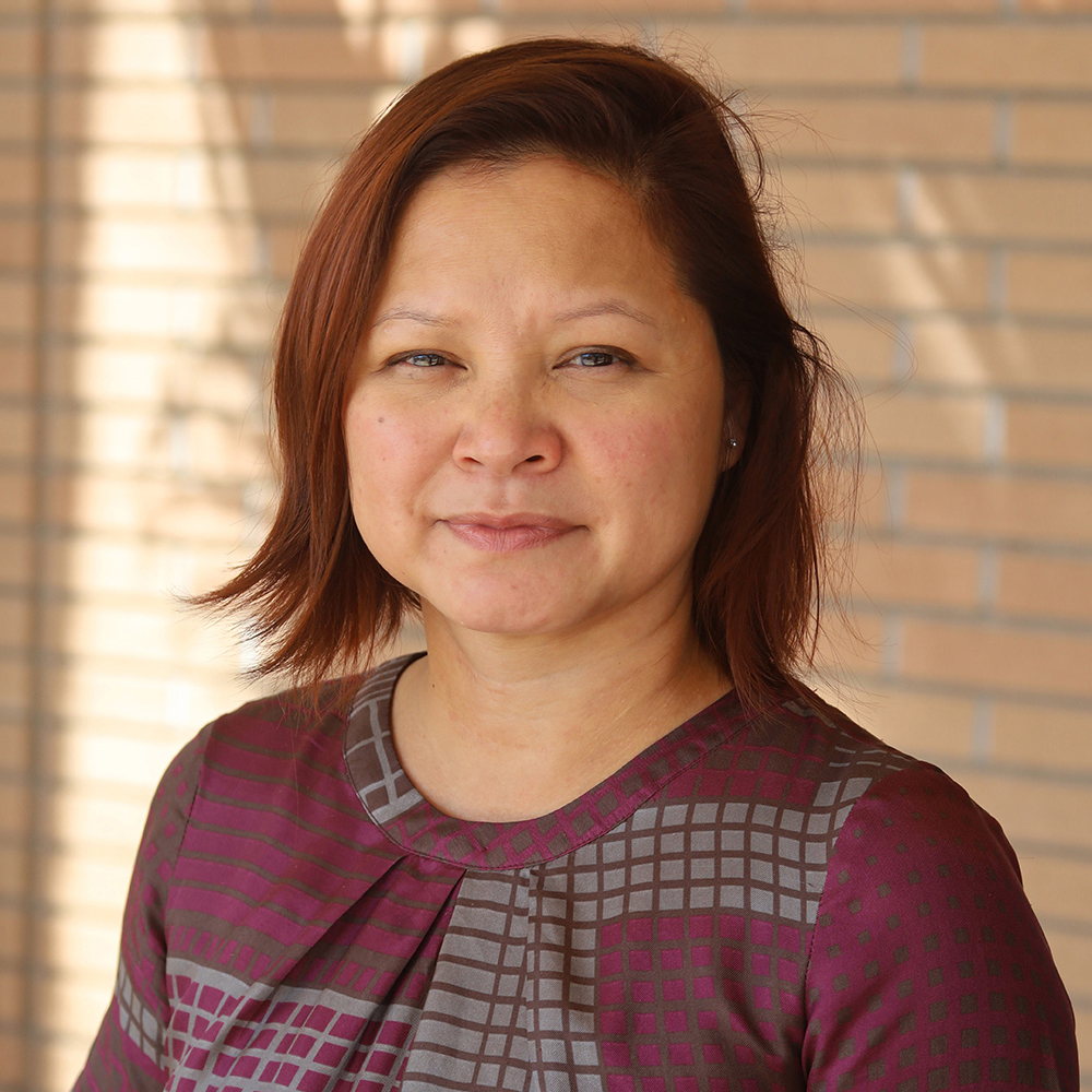 Nhung Tuyet Tran Color photograph of an Asian woman with auburn hair wearing a purple and gray gridded top; she stands in front of a brick wall and looks at the camera with a slight smile