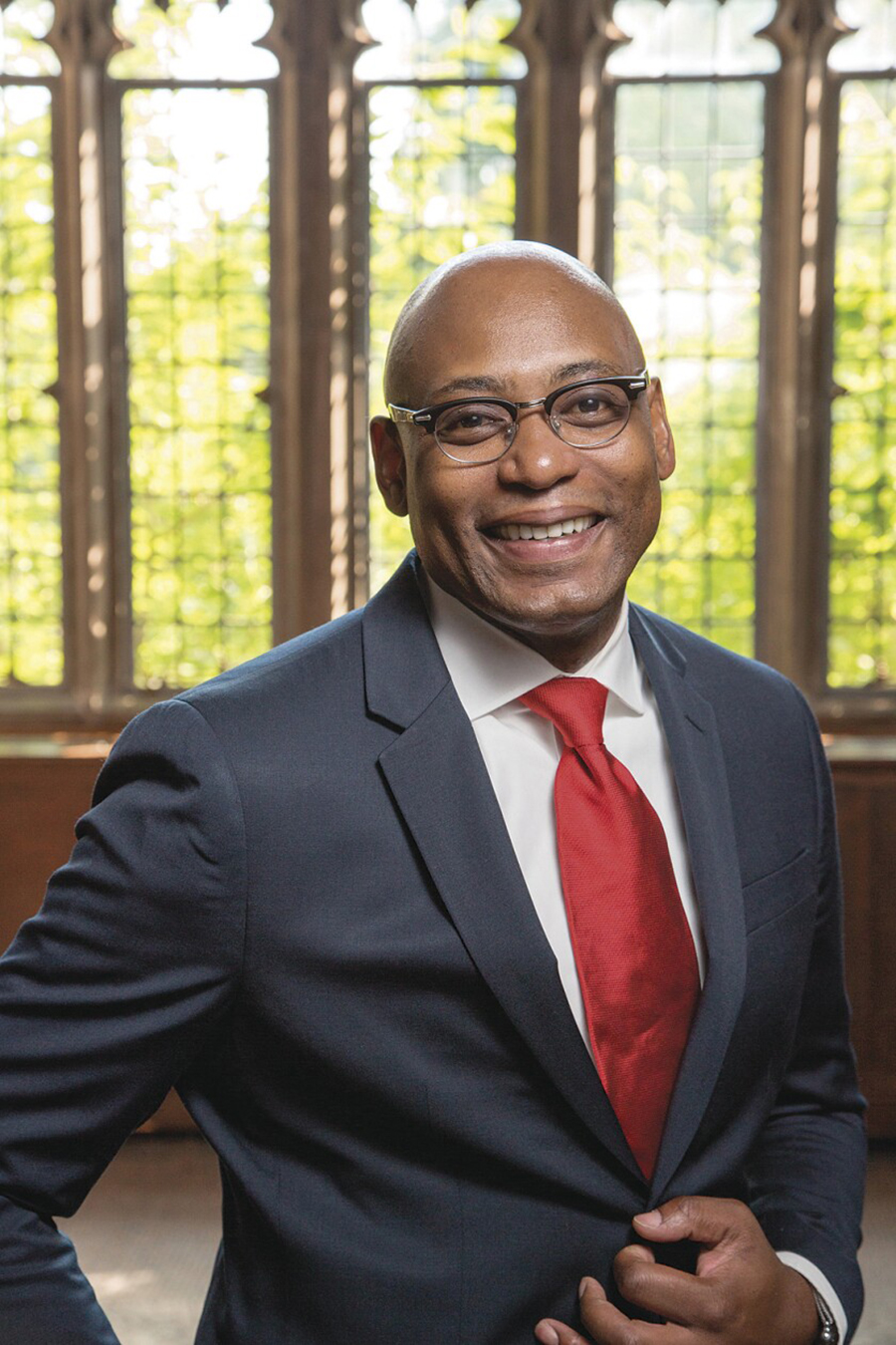 Patrice Rankine Color photograph of a dark skinned man wearing a suit and tie; he stands in front of stained glass windows and smiles at the camera