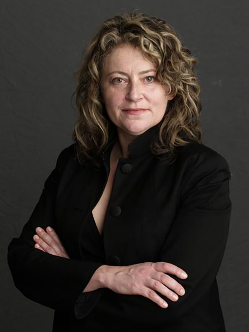 Photo of a light skinned woman with wavy hair and her arms crossed, looking at the camera