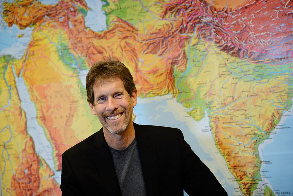 Resident - Kenneth Baxter Wolf Color photograph of a light skinned man with brown hair smiling at the camera; he wears a dark blazer and is positioned in front of a large world map that spans from Egypt on the left to India on the right