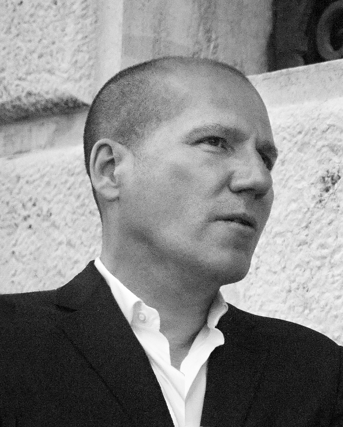 Resident - Michael Rock Black and white photograph of the head and shoulders of a light skinned man with close cropped hair wearing a dark blazer over a white shirt; he is shown in 3/4 profile looking to the right side