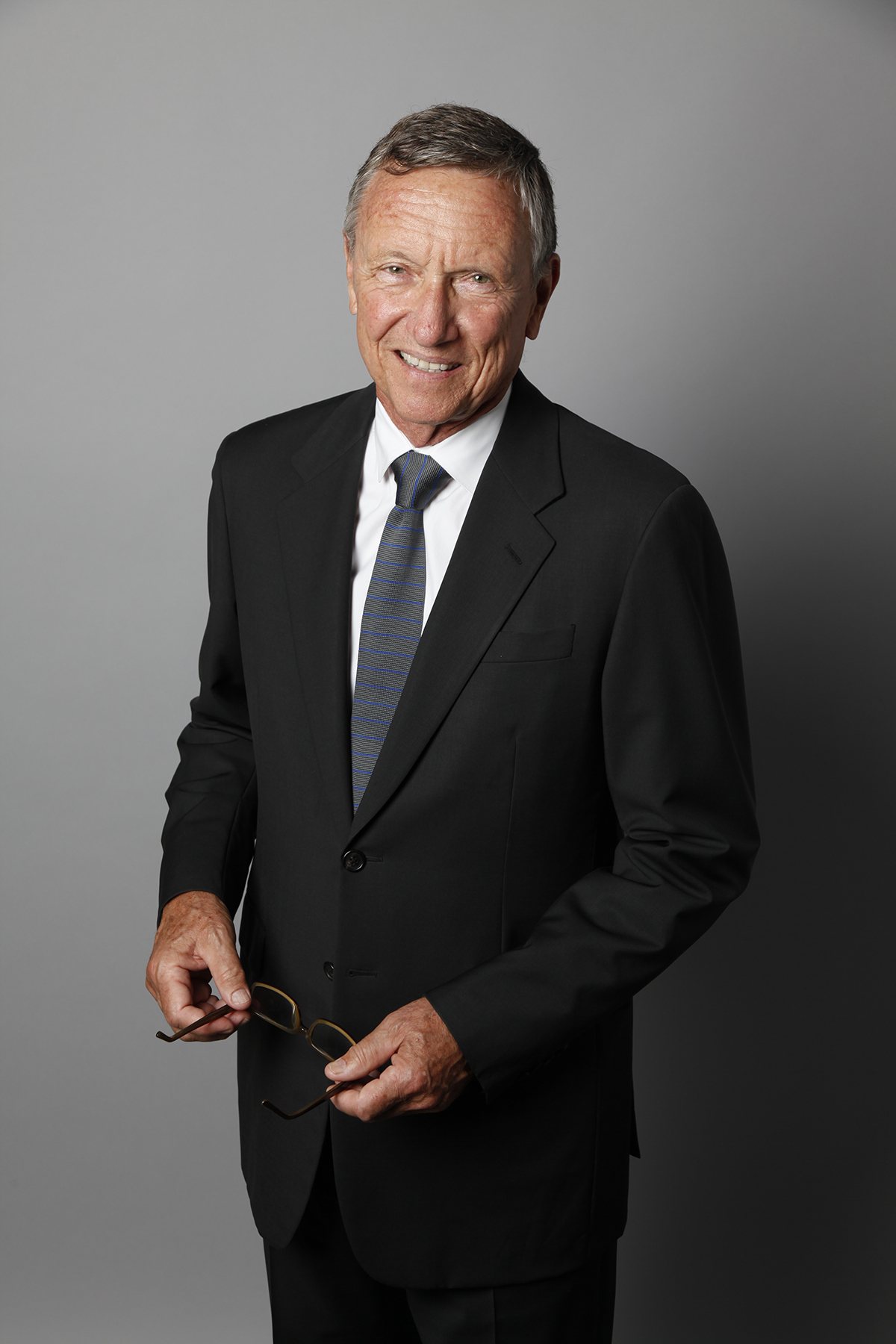Richard Gluckman Color photograph of a light skinned man wearing a dark suit and tie standing against a neutral gray backdrop and smiling at the camera