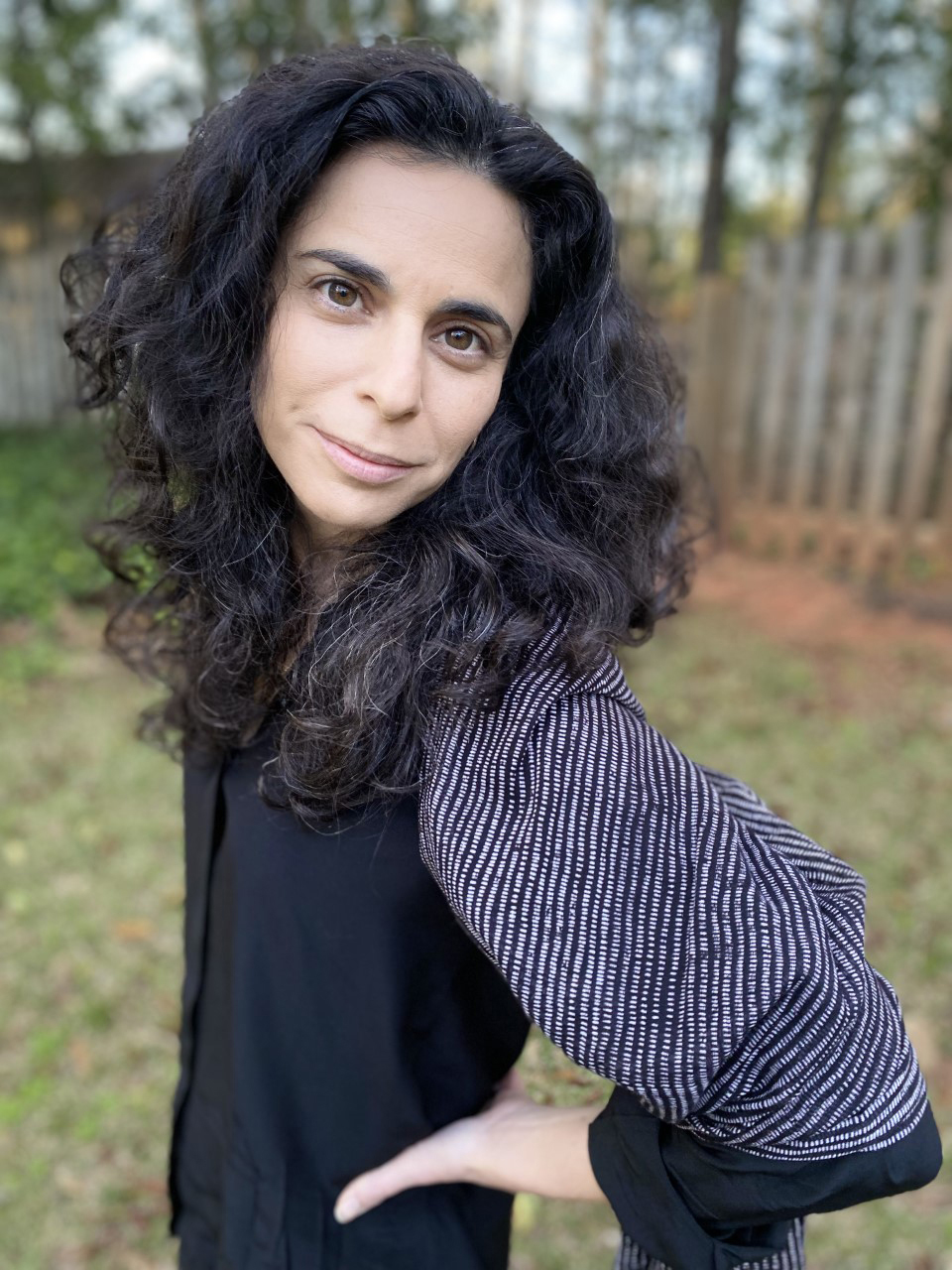 Robyn Schiff Color photograph of a light skinned woman with dark wavy hair standing in a fenced-in backyard; she stands facing left with her hands on her hips, looking directly into the camera