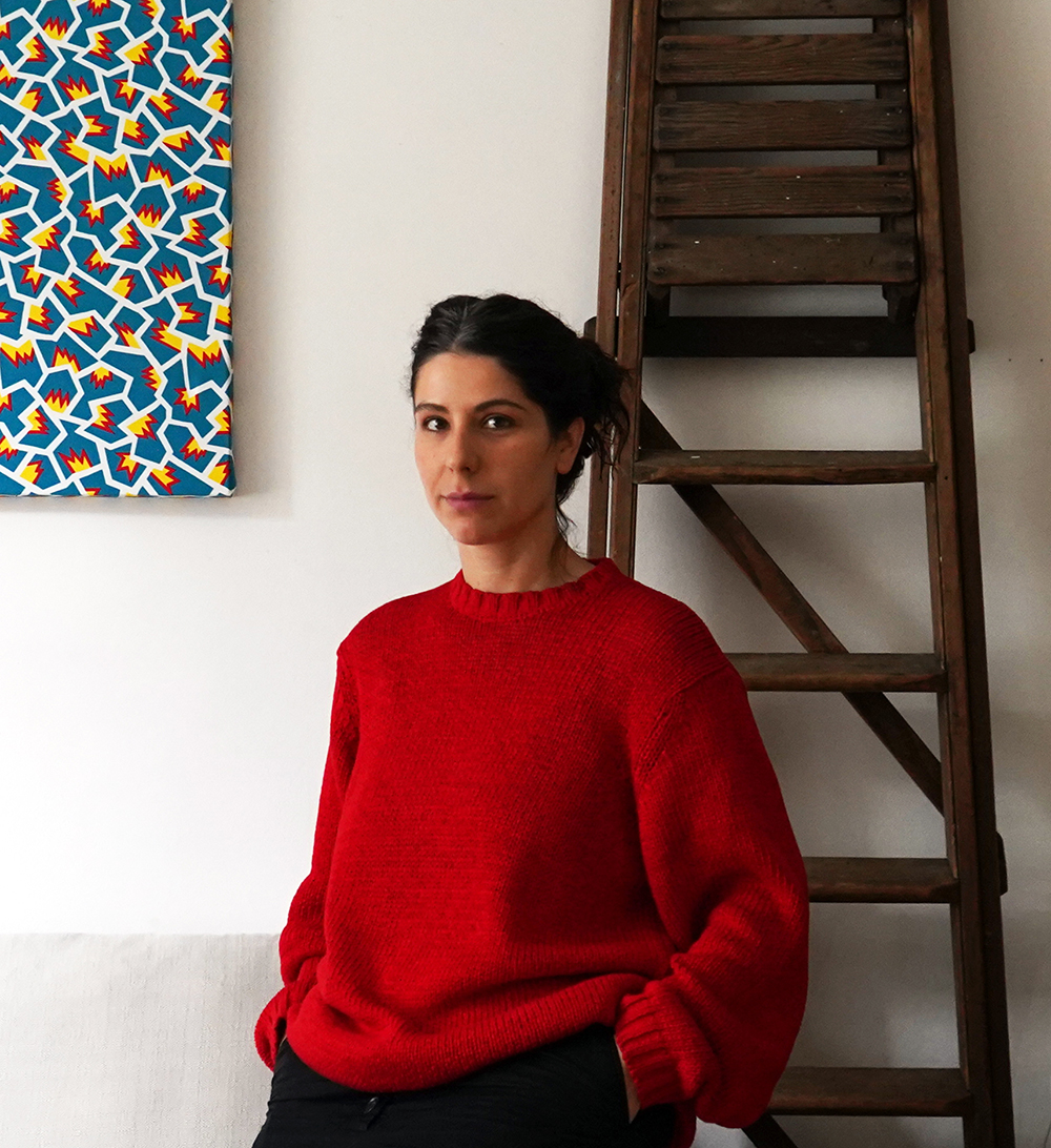 Sabrina Morreale Color photograph of a light skinned woman wearing a red sweater; she stands indoors next to a ladder leaning on the wall, looking directly at the camera