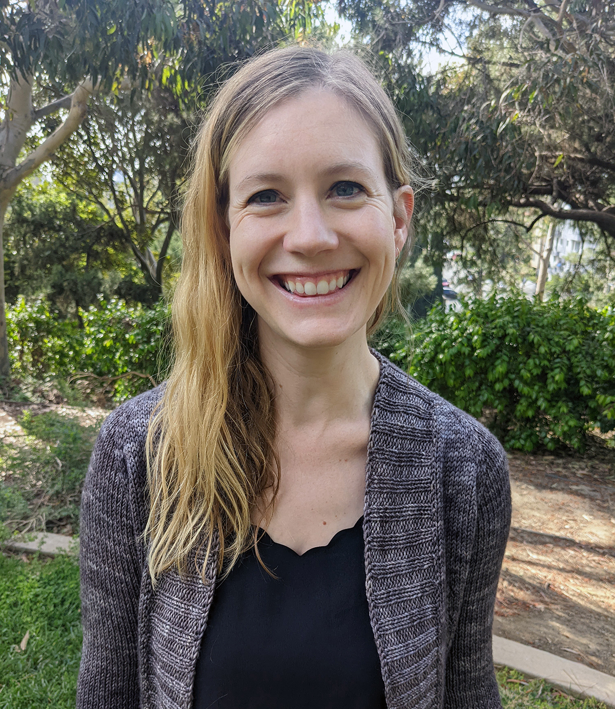 Sarah Beckmann Color photograph of the head and torso of a light skinned woman with long blond hair standing in an outdoor garden location; she smiles directly at the camera