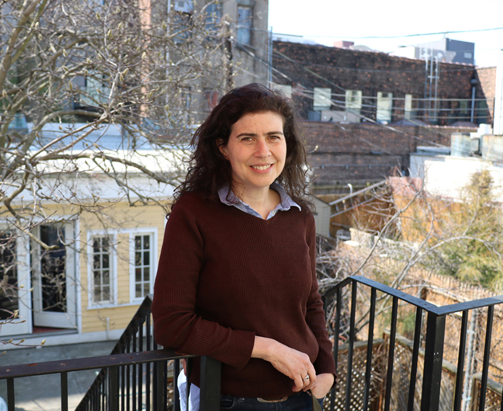 Sarah Nunberg Color photograph of Sarah Nunberg standing on a patio with several New York building backyards behind her