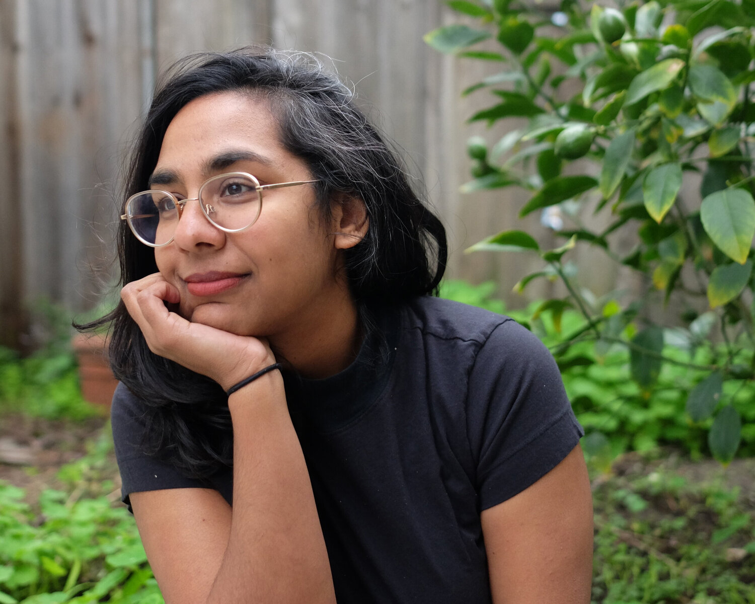 Shruti Swamy Color photograph of a brown skinned woman with dark hair wearing glasses; she sits in a garden or backyard location with a wooden fence behind her, her hand resting on her chin as she looks off camera