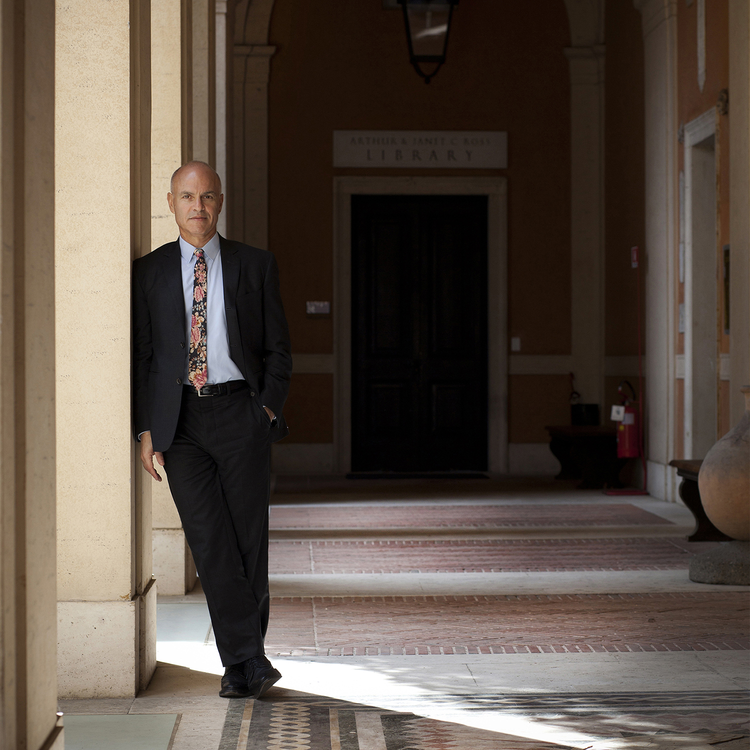 Staff - Mark Robbins Library Color photograph of a light skinned man wearing a dark suit and leaning against a column in a Roman cortile