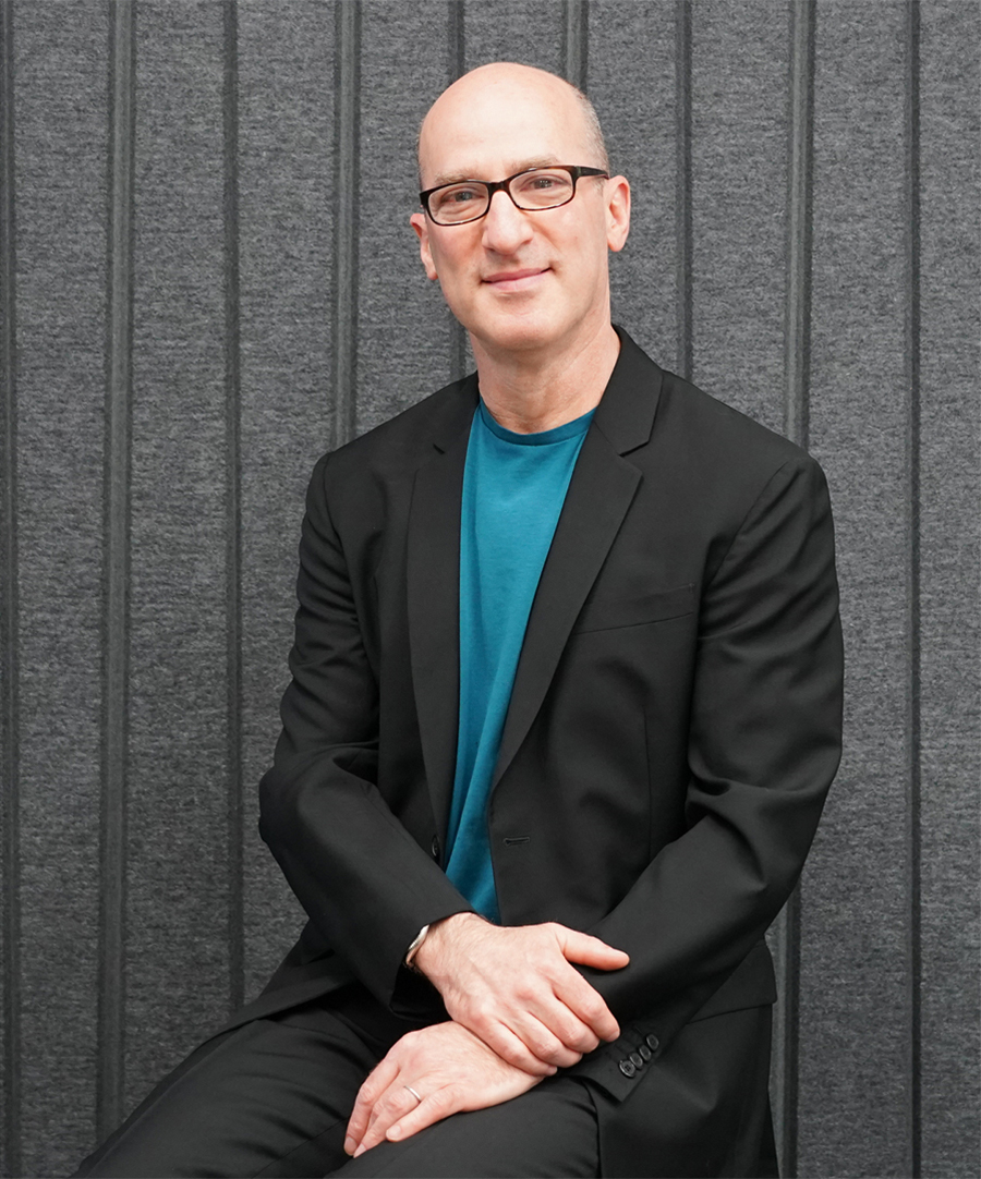 Stephen Cassell Color photograph of a light skinned man seen from the waist up looking at the camera; he wears glasses and an aqua shirt under a dark blazer