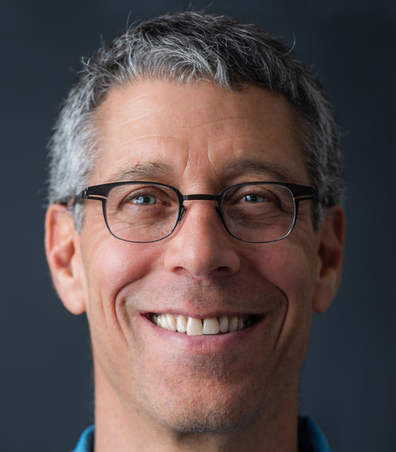 Color photograph of the face of a light skinned man with gray and dark hair, wearing glasses and smiling confidently into the camera