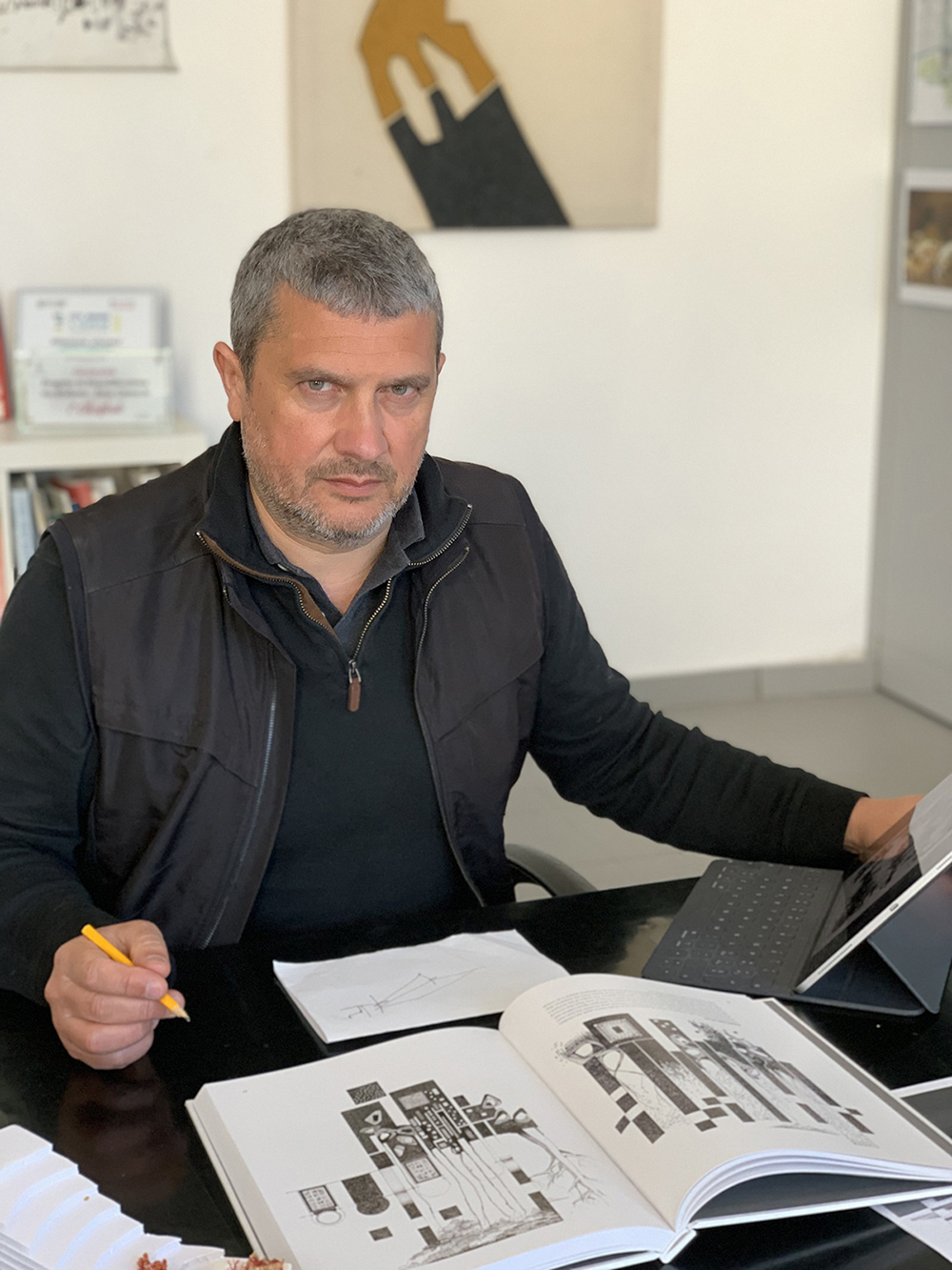 Valerio Morabito Color photograph of Valerio Morabito sitting at a table with a pencil in hand and a drawing on paper, an open book, and a tablet computer in front of him