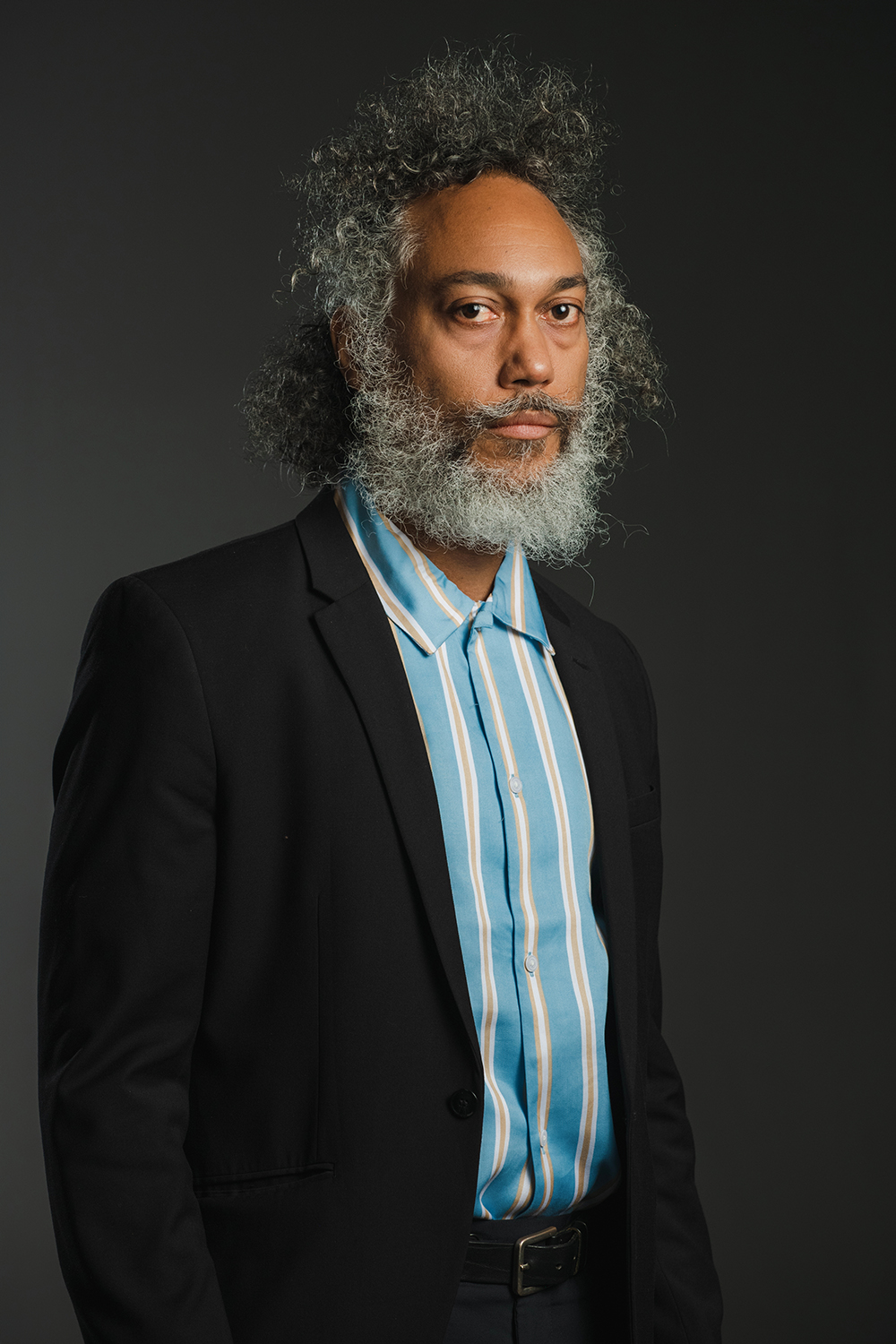 Zachary Fabri Color photograph of a dark skinned man with gray hair and beard wearing a dark blazer over a blue shirt; he looks at the camera with a serious expression