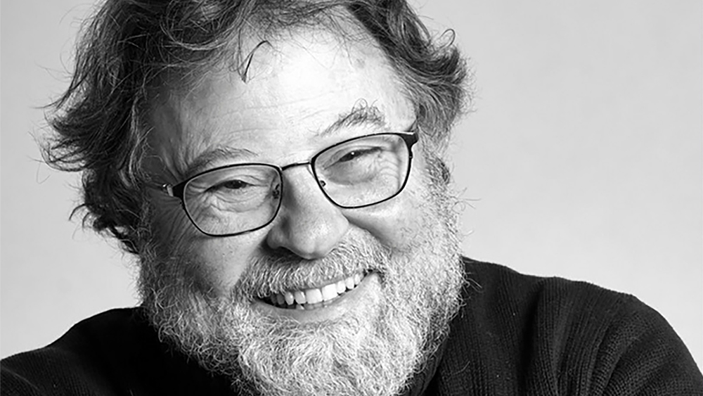Black and white photo of the head and shoulders of a light skinned man with a beard, glasses, and a big grin