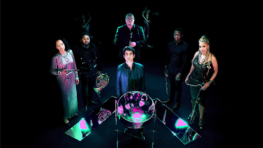 Color photo of six musicians standing in the dark look up at the camera